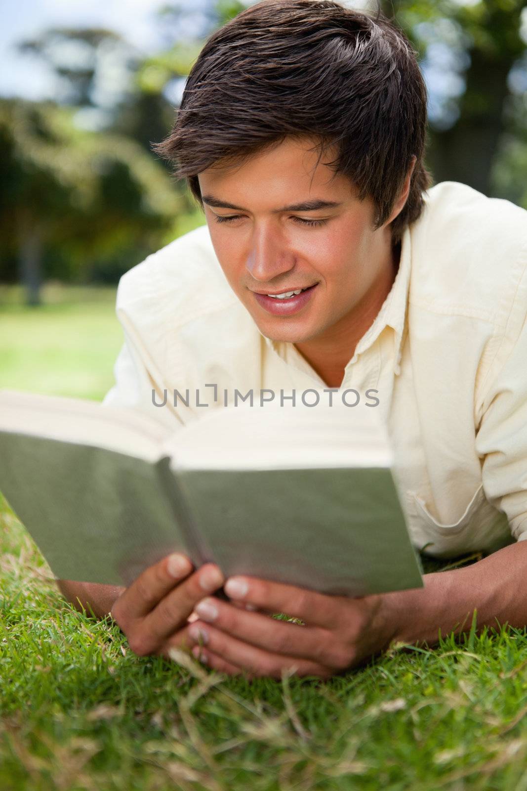 Man smiles while he reads a book as he lies prone on the grass