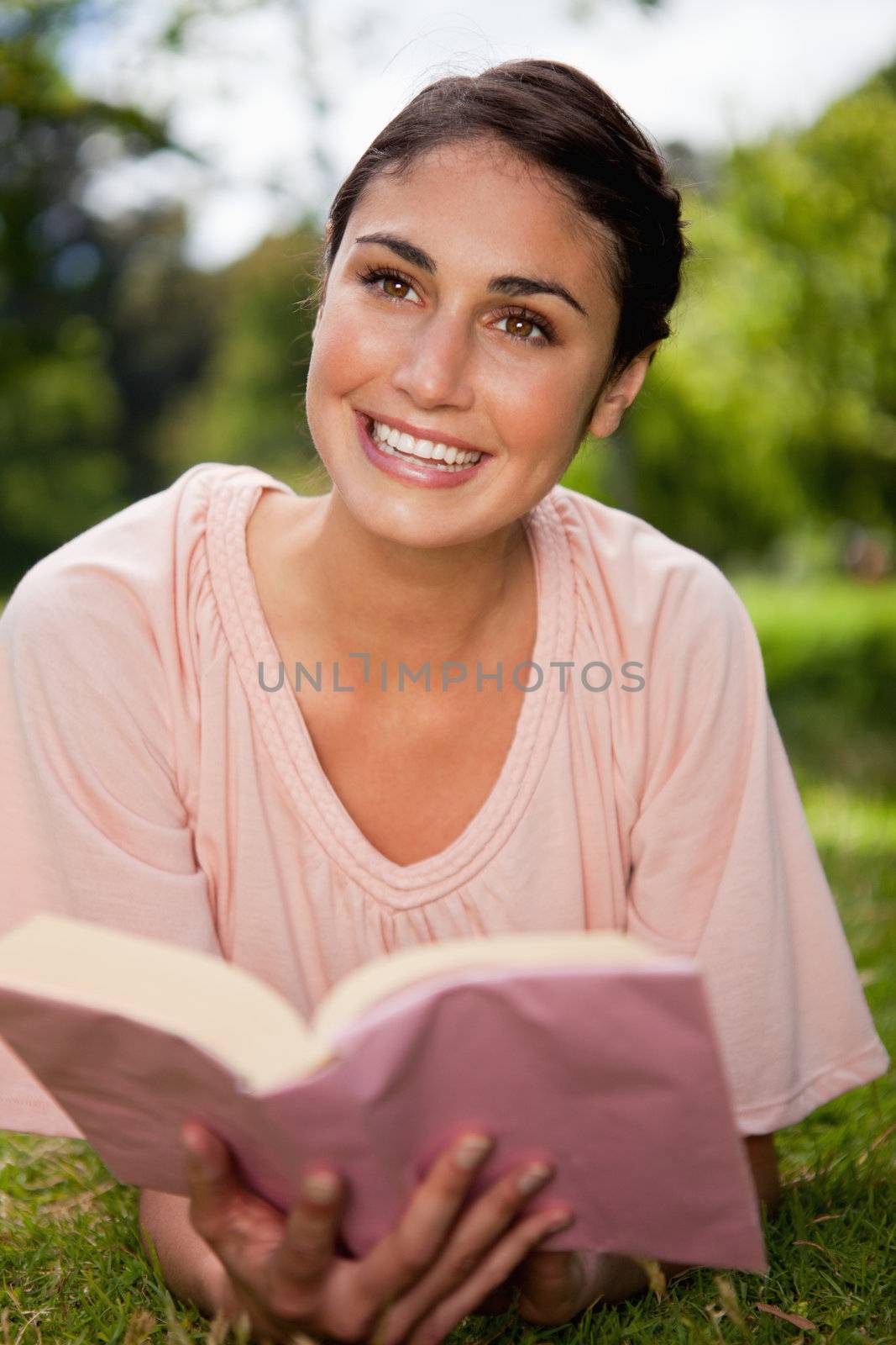 Woman looks towards to the sky while she reads a books as she is lying prone on grass