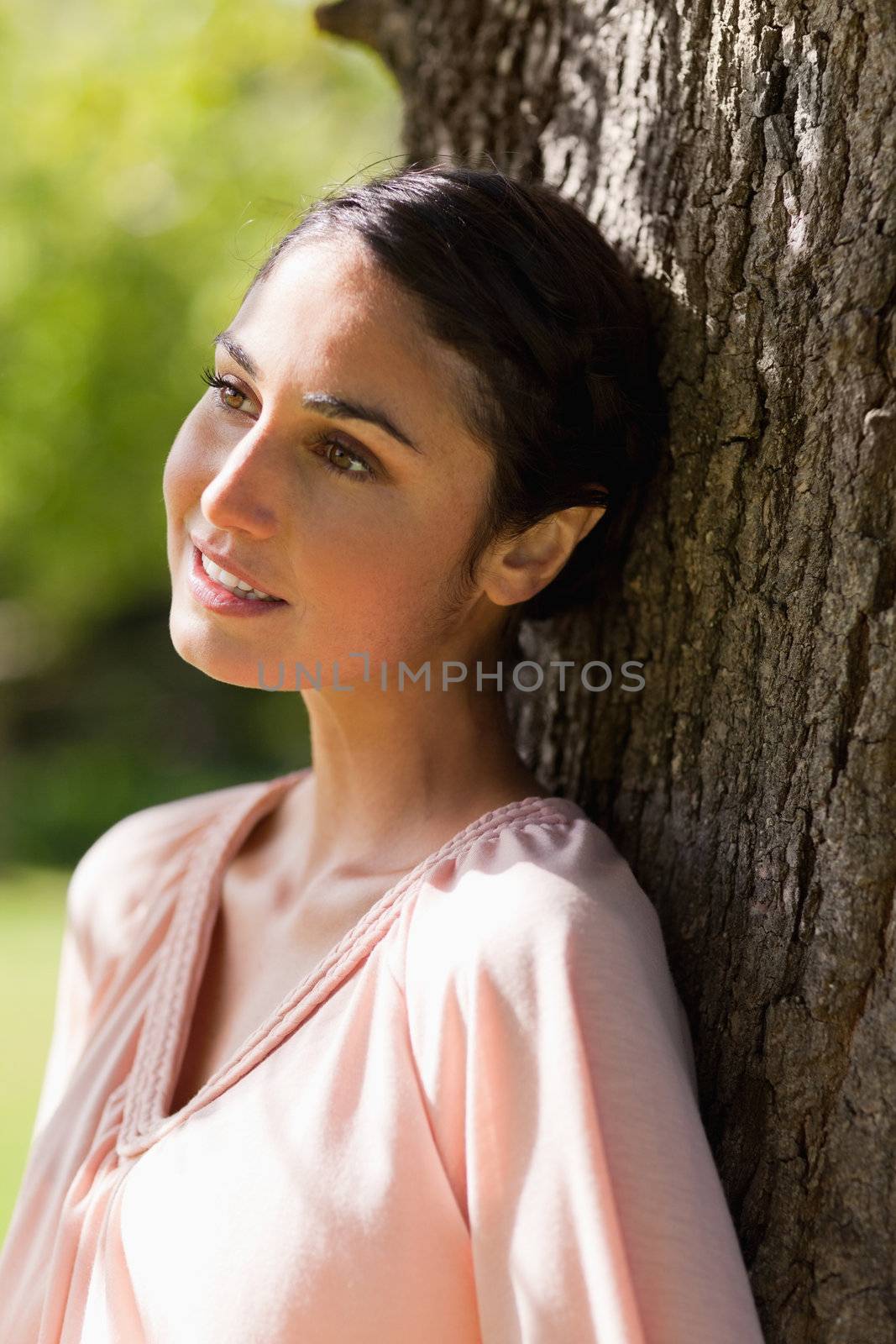 Young woman looking ahead of her while and smiling sitting against the trunk of a tree