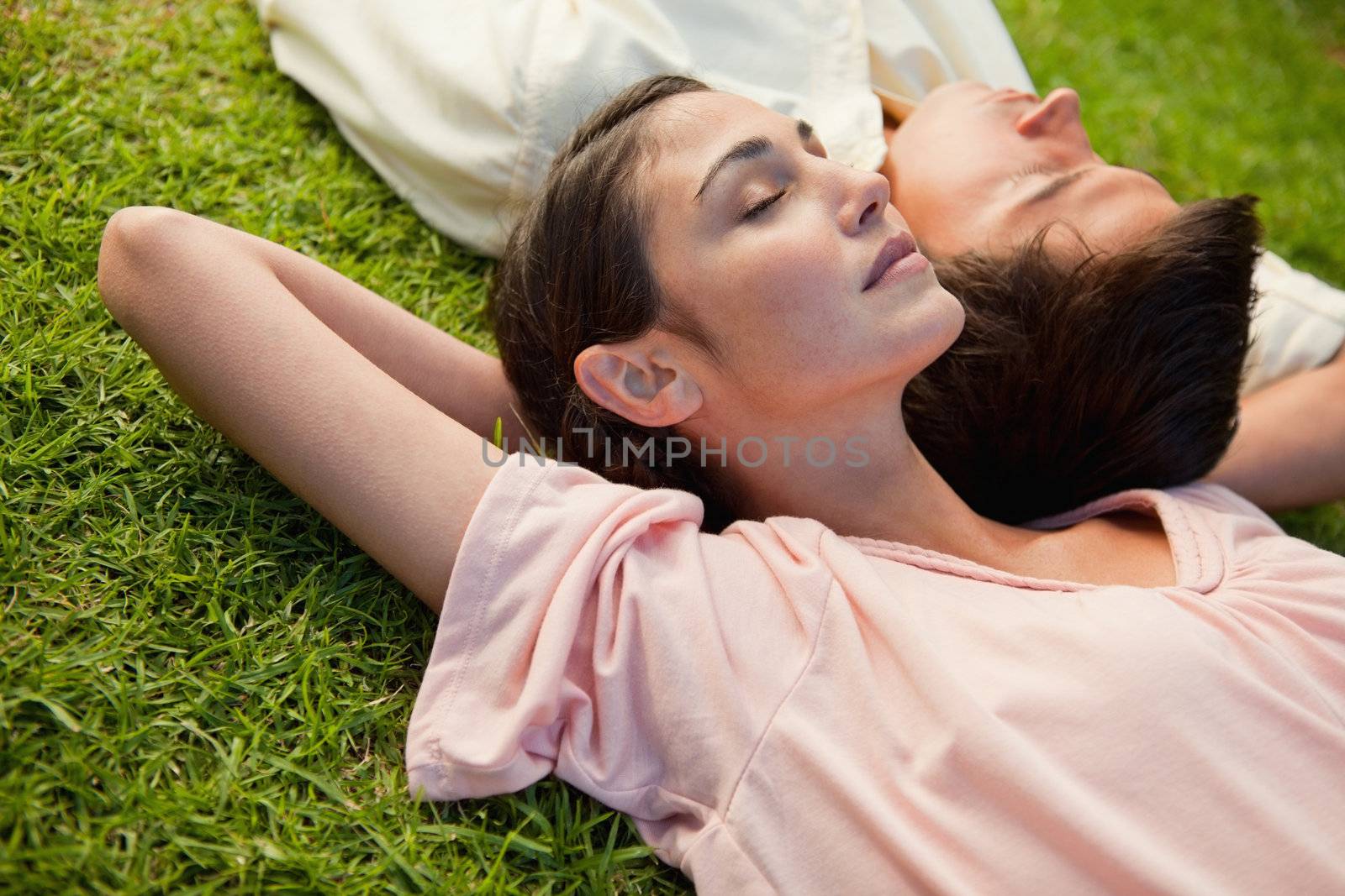 Woman and a man with their eyes closed lying head to shoulder with their arms resting behind their neck on the grass