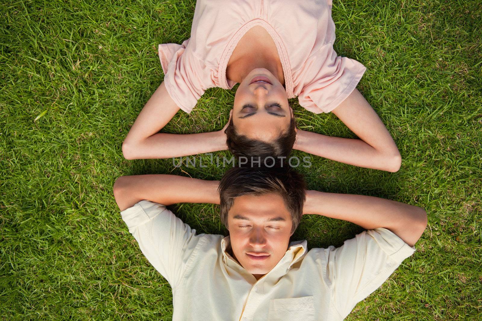 Man and a woman with their eyes closed lying head to head with both of their arms resting behind their neck on the grass