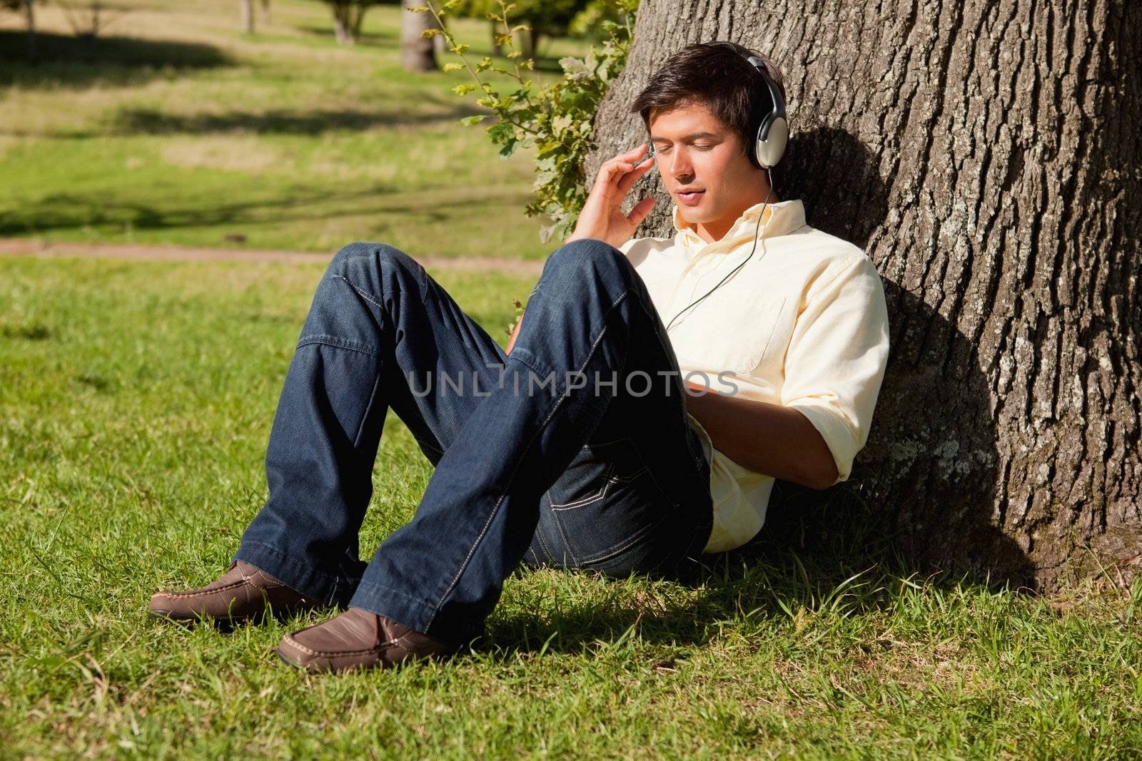 Man using headphones to listen to music while resting against the trunk of a tree