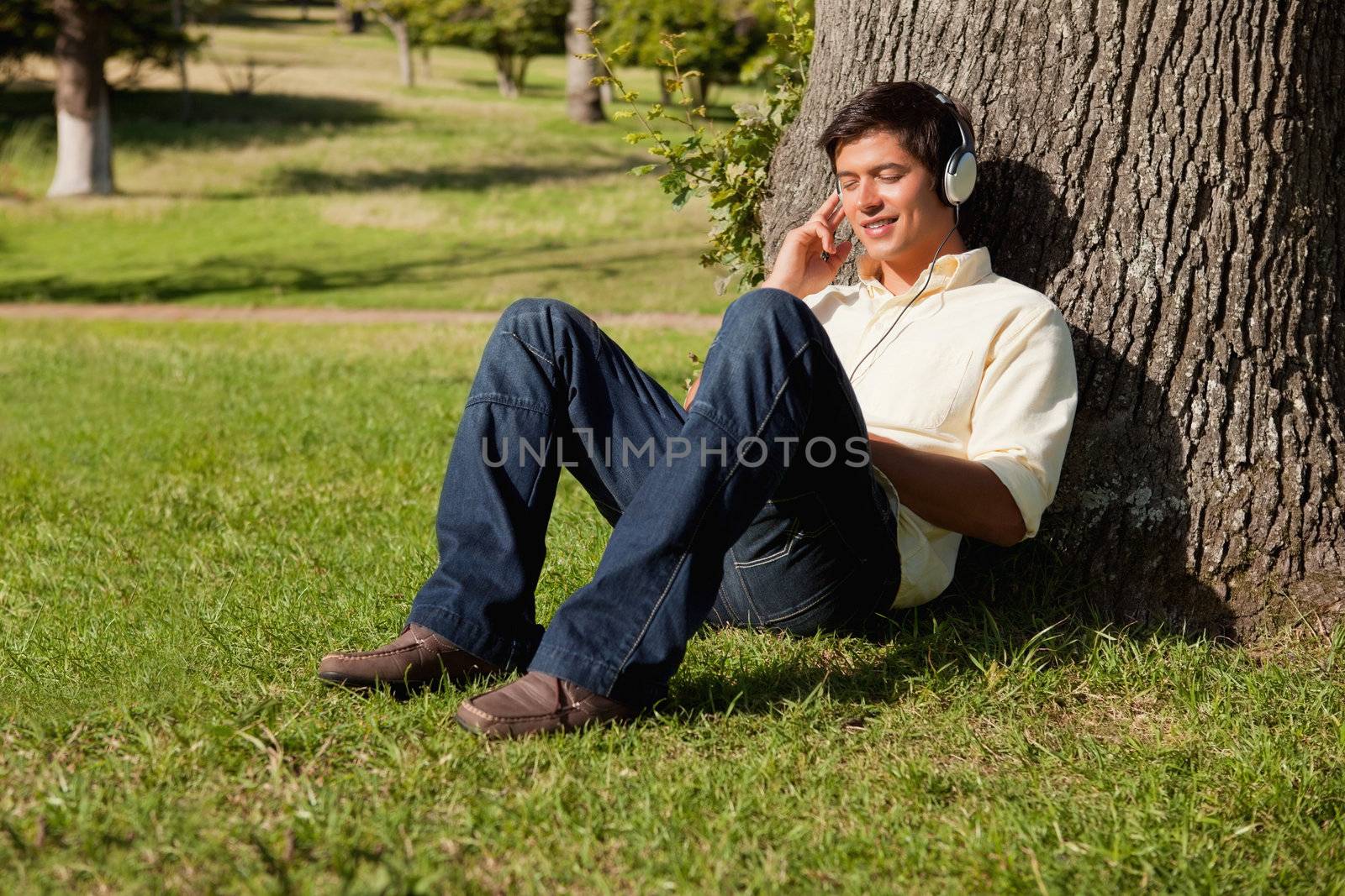 Man smiling as he uses headphones to listen to music while resting against the trunk of a tree
