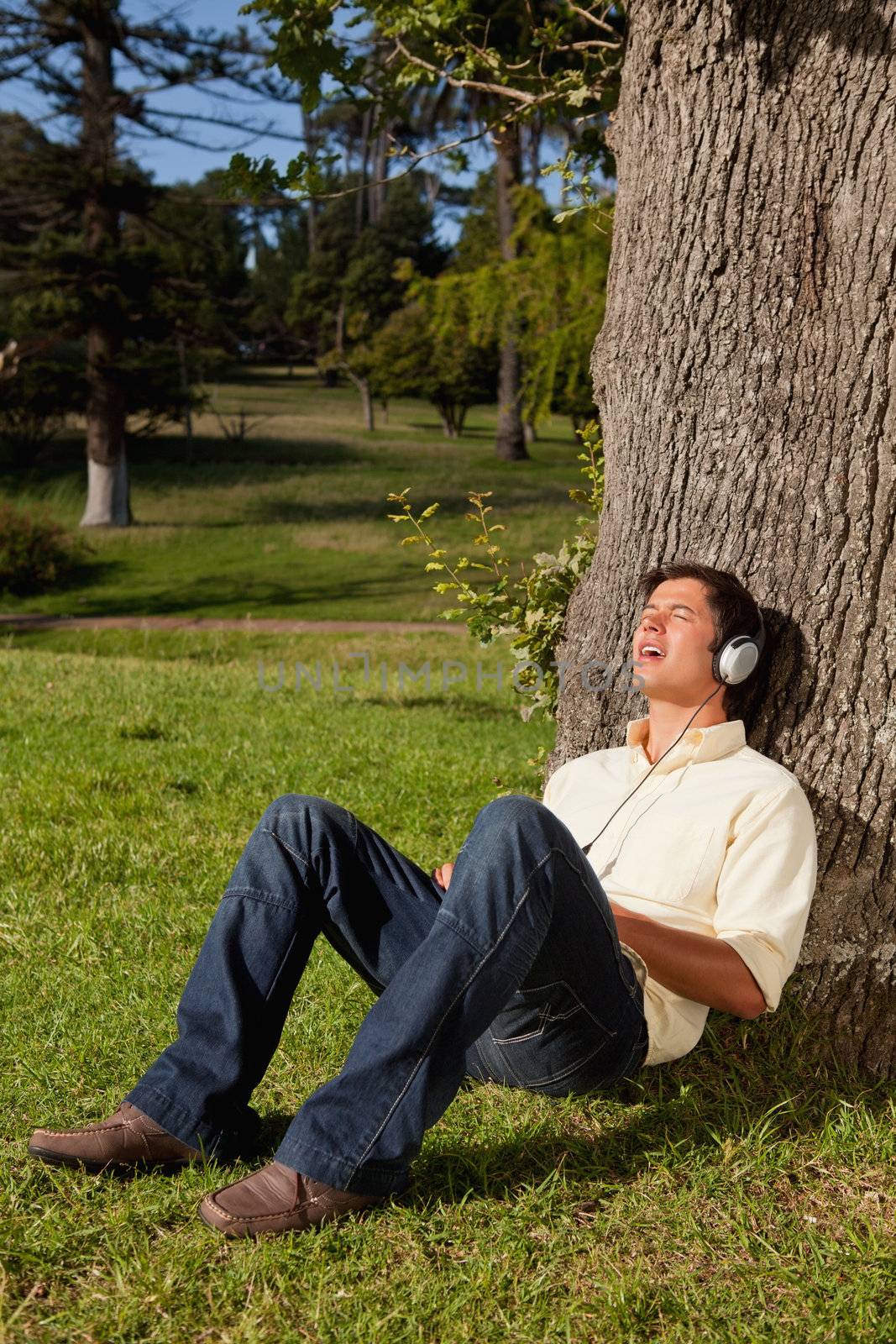 Man singing while using headphones to listen to music as he rests against the trunk of a tree