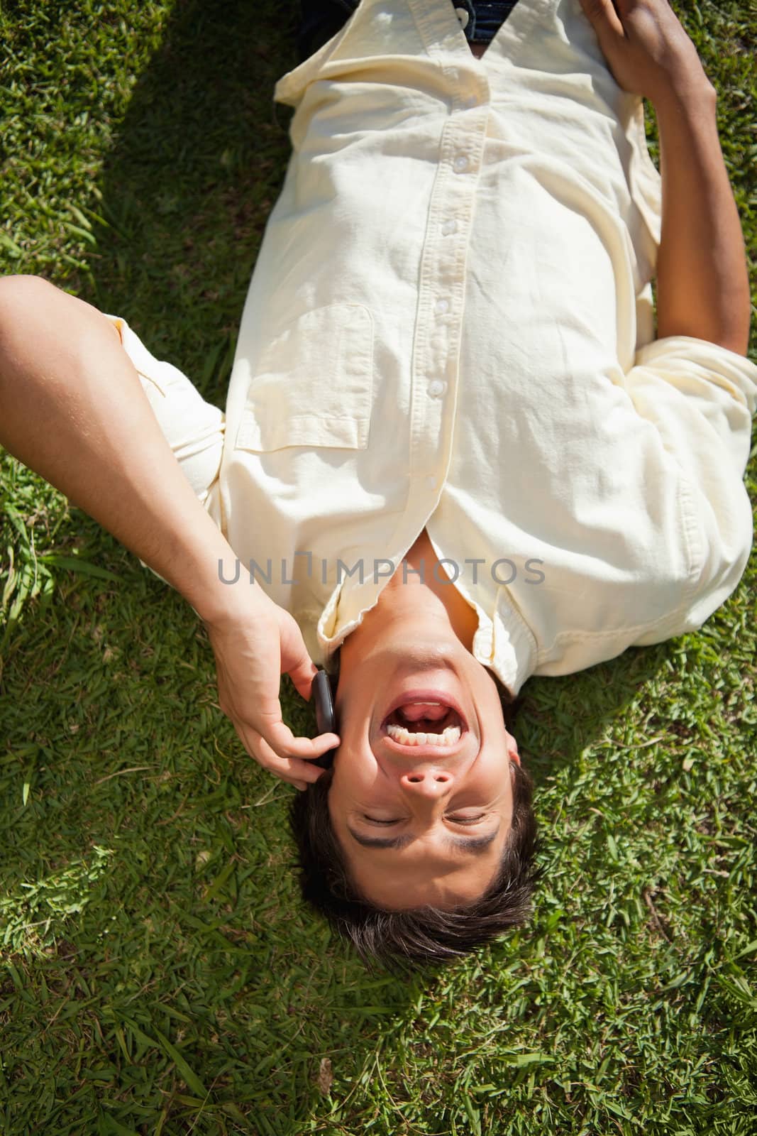 Man with his eyes closed laughing while using a mobile phone as he lies down on the grass