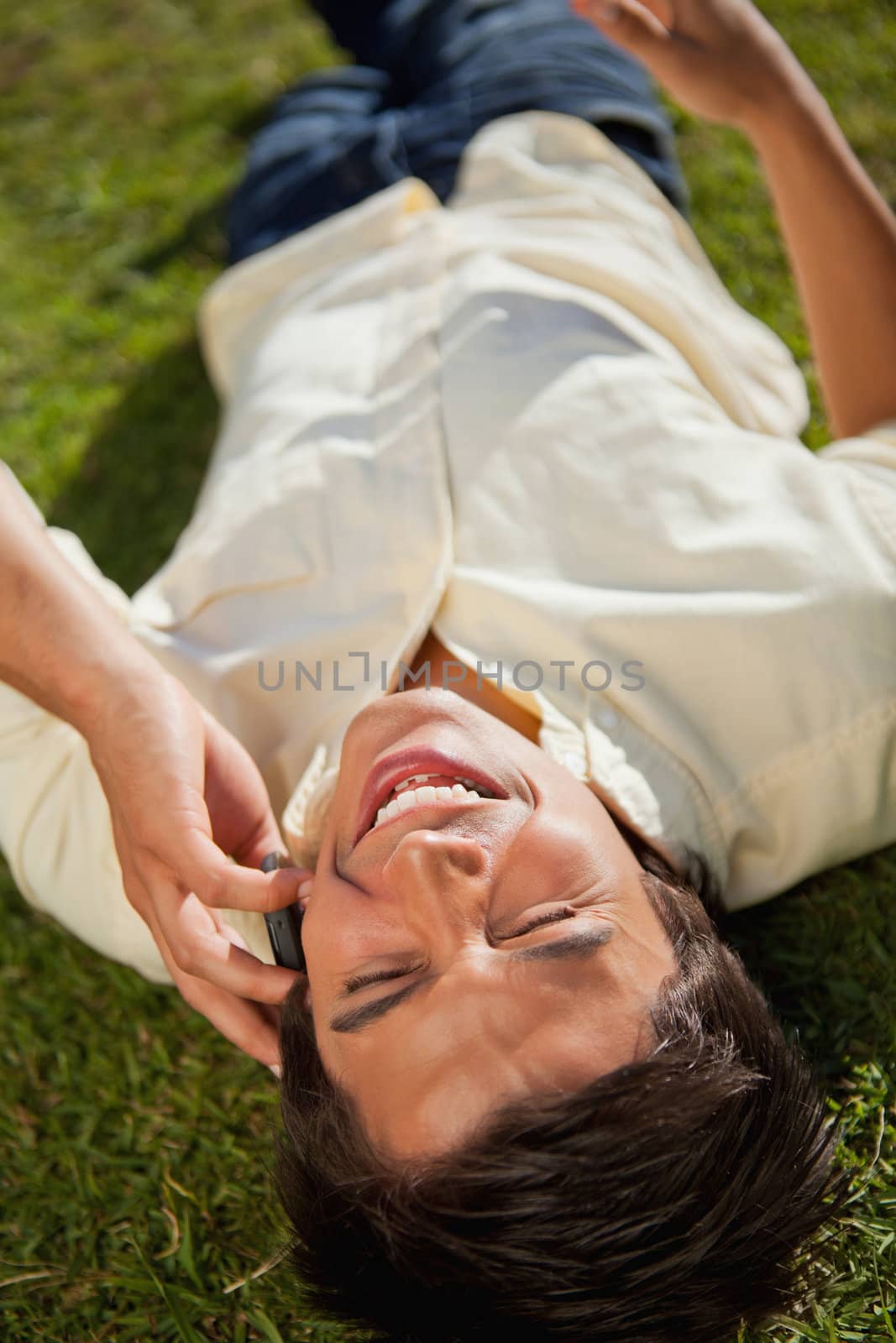 Close-up of a man with his eyes closed laughing while making a call over the phone as he lies down on the grass
