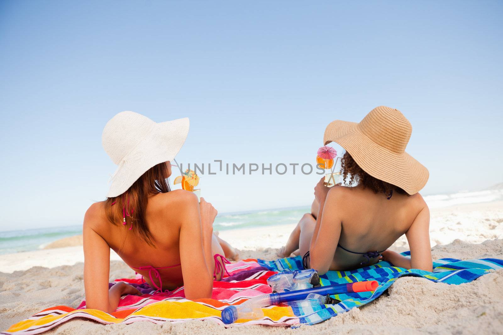 Back view of beautiful women sunbathing on beach towels while sipping cocktails