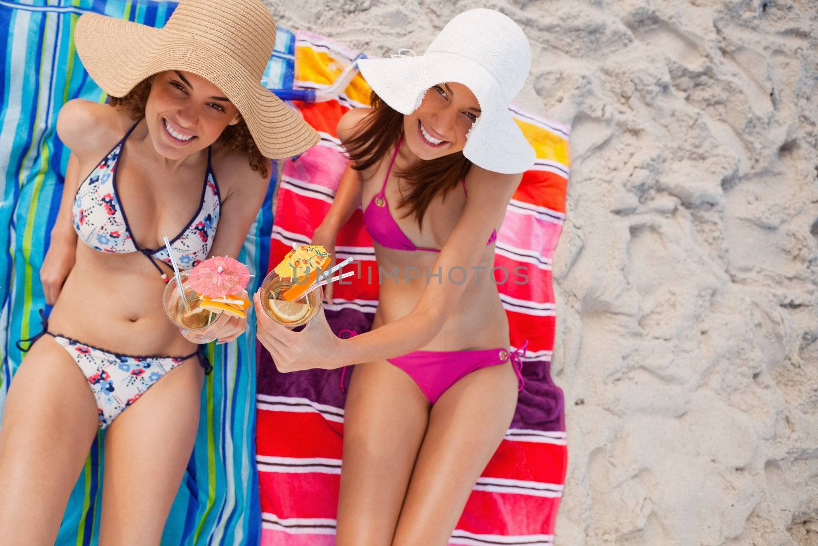 Beautiful women raising their cocktails together while sitting on beach towels