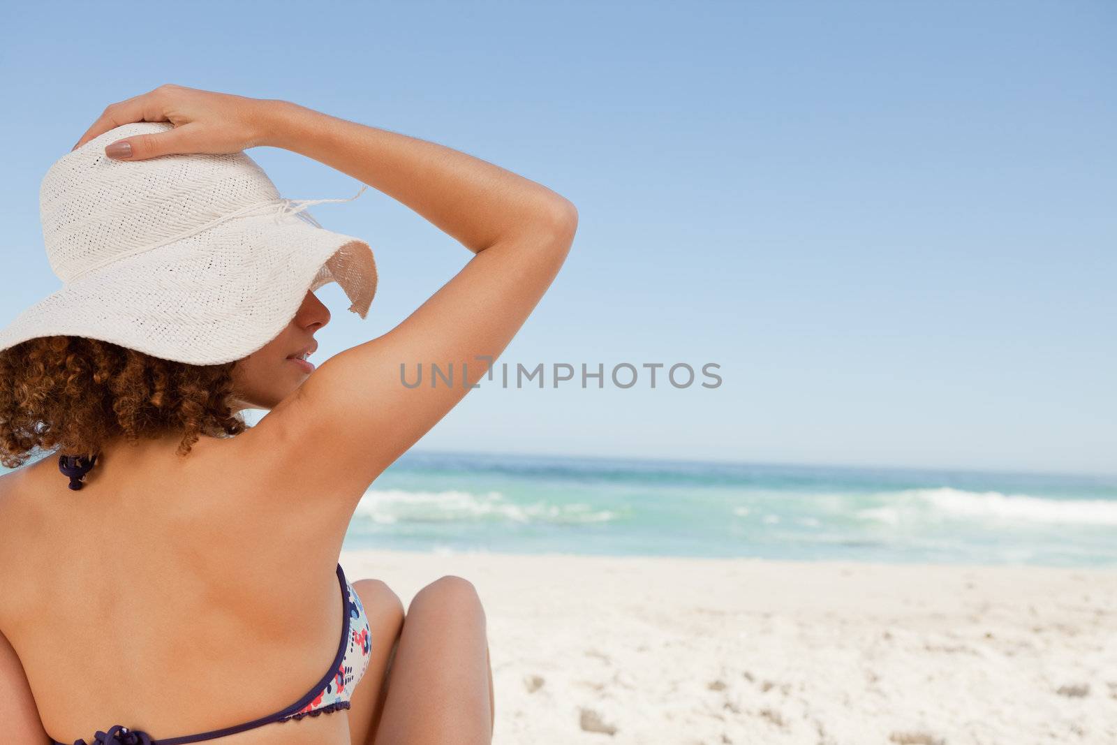 Young woman sitting in front of the ocean while holding her hat on her head