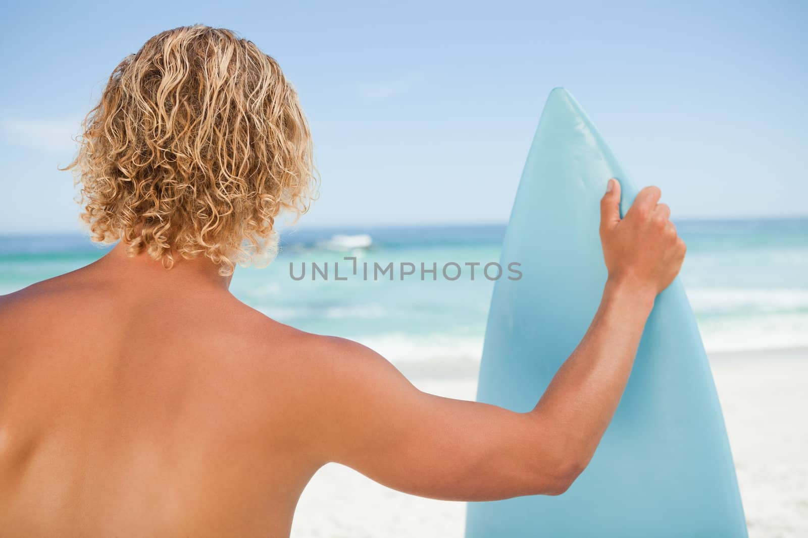 A young blonde man holding a perched surfboard while standing on the beach