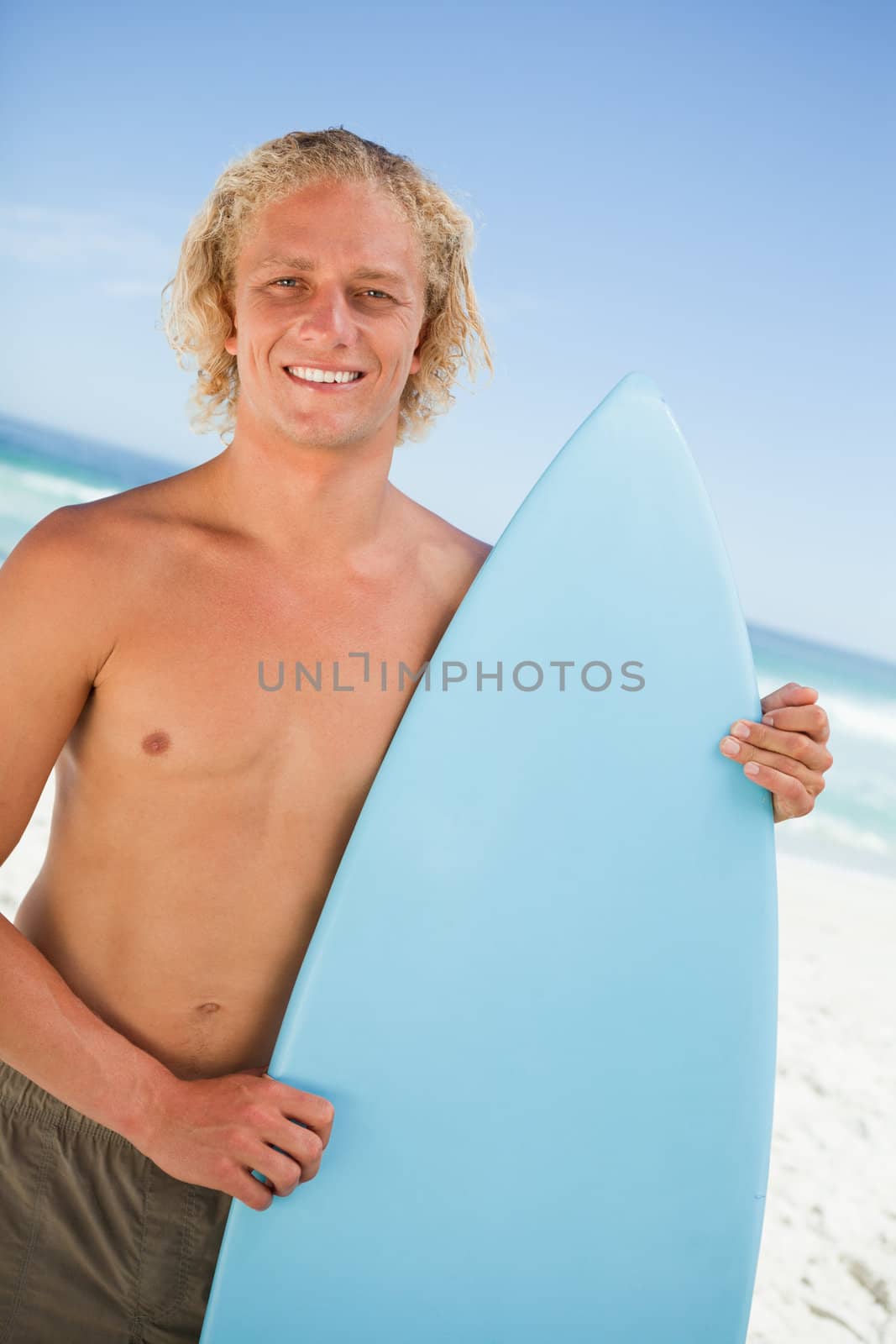 Young smiling man standing upright on the beach while holding his surfboard