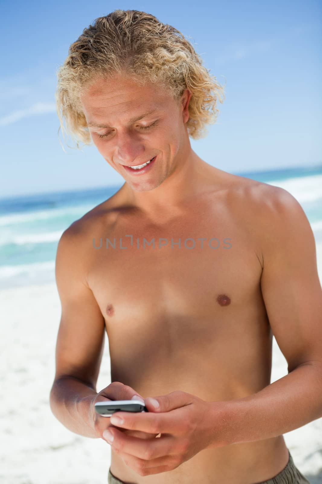 Smiling blonde man standing on the beach while sending a text  by Wavebreakmedia