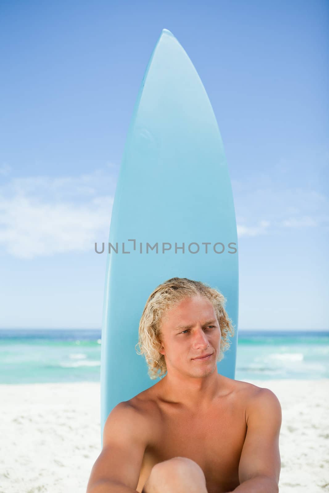 Serious blonde man sitting on the beach in front of his blue surfboard