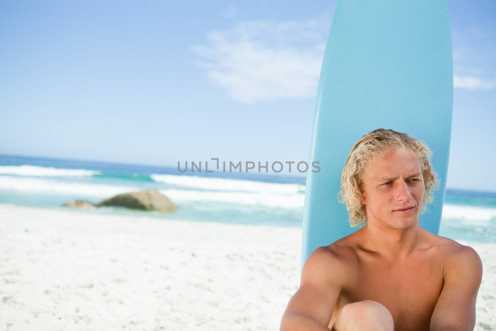 Blonde man sitting on the beach with his surfboard while looking towards the side