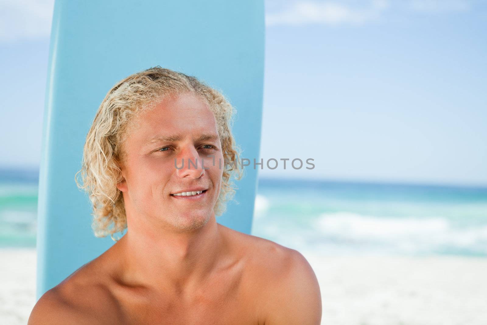 Young blonde man sitting on the beach with his surfboard while showing a smile