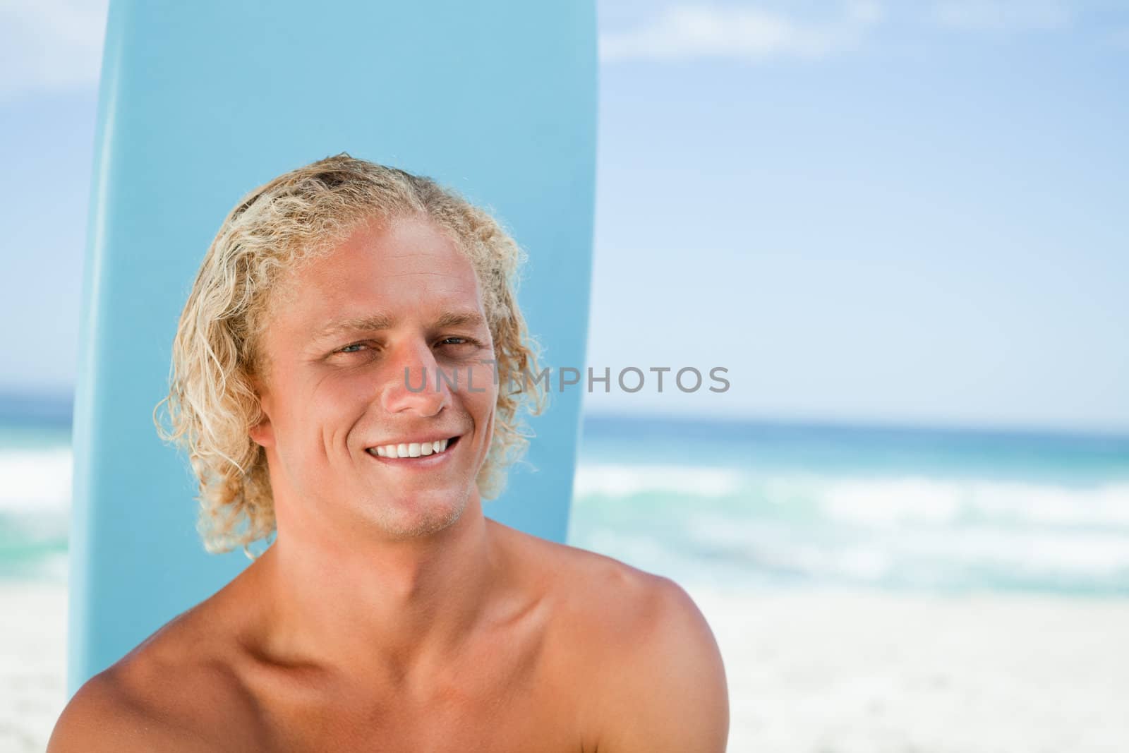Young man sitting in front of the ocean with his blue surfboard while smiling