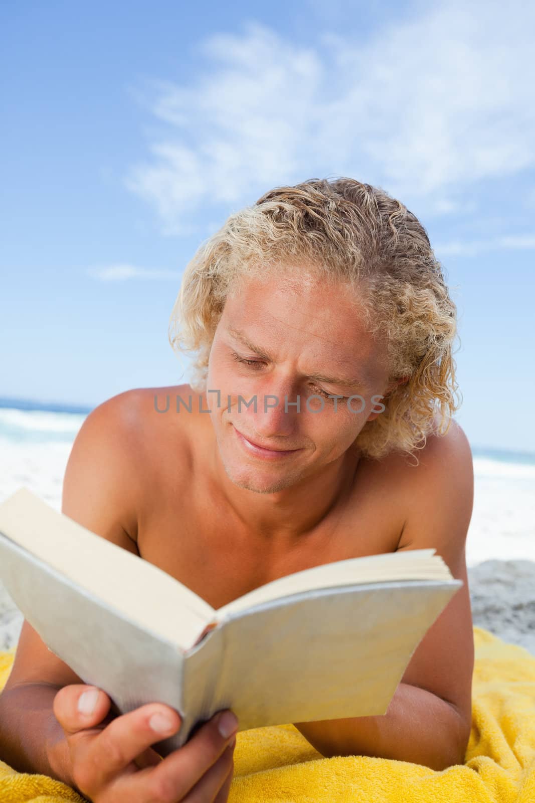 Young blonde man lying on his beach towel while seriously reading a book