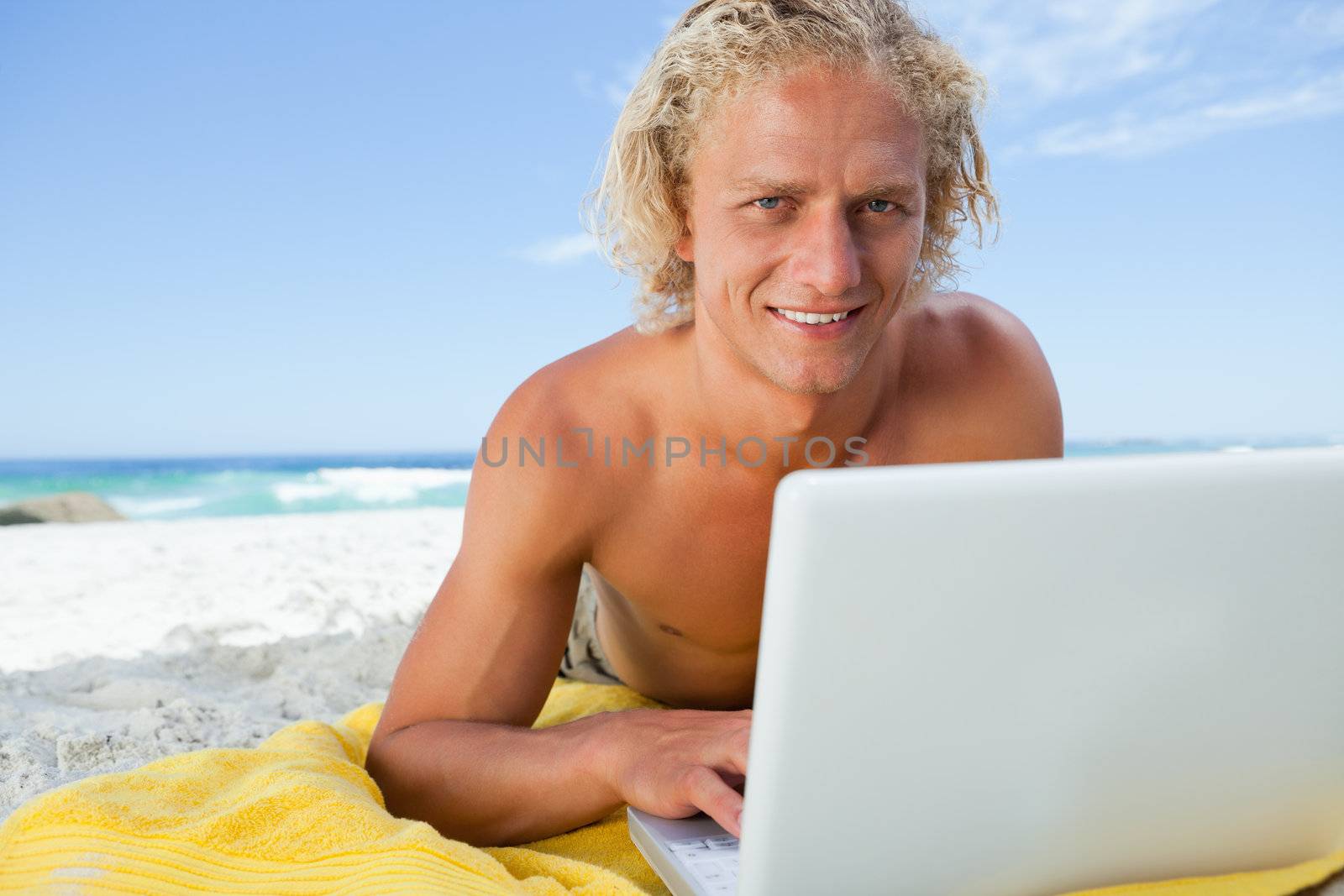 Blonde man lying on the beach while using his laptop and showing a great smile