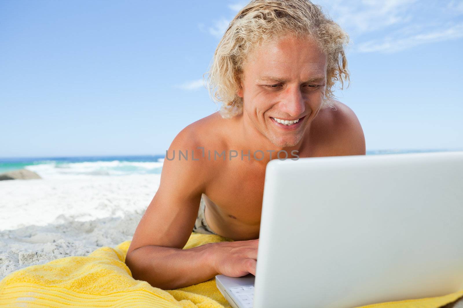 Smiling blonde man looking at his laptop while lying on the beach by Wavebreakmedia