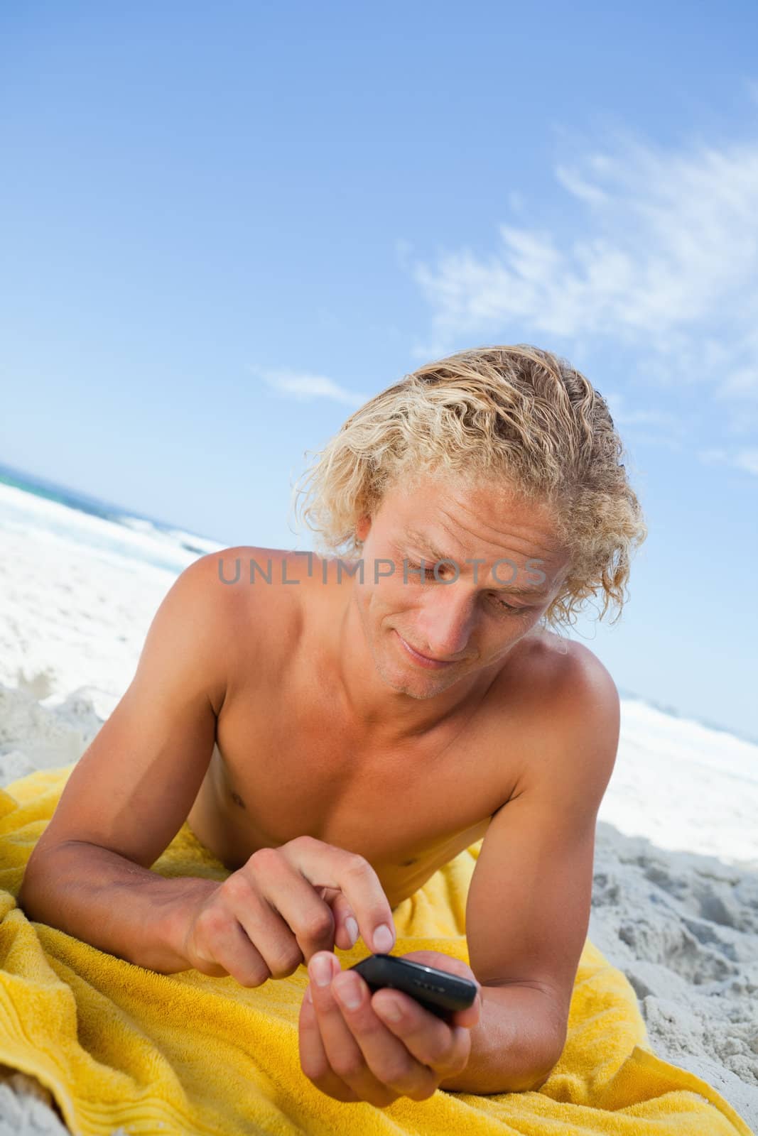 Young blonde man sending a text with his cellphone while lying on the beach