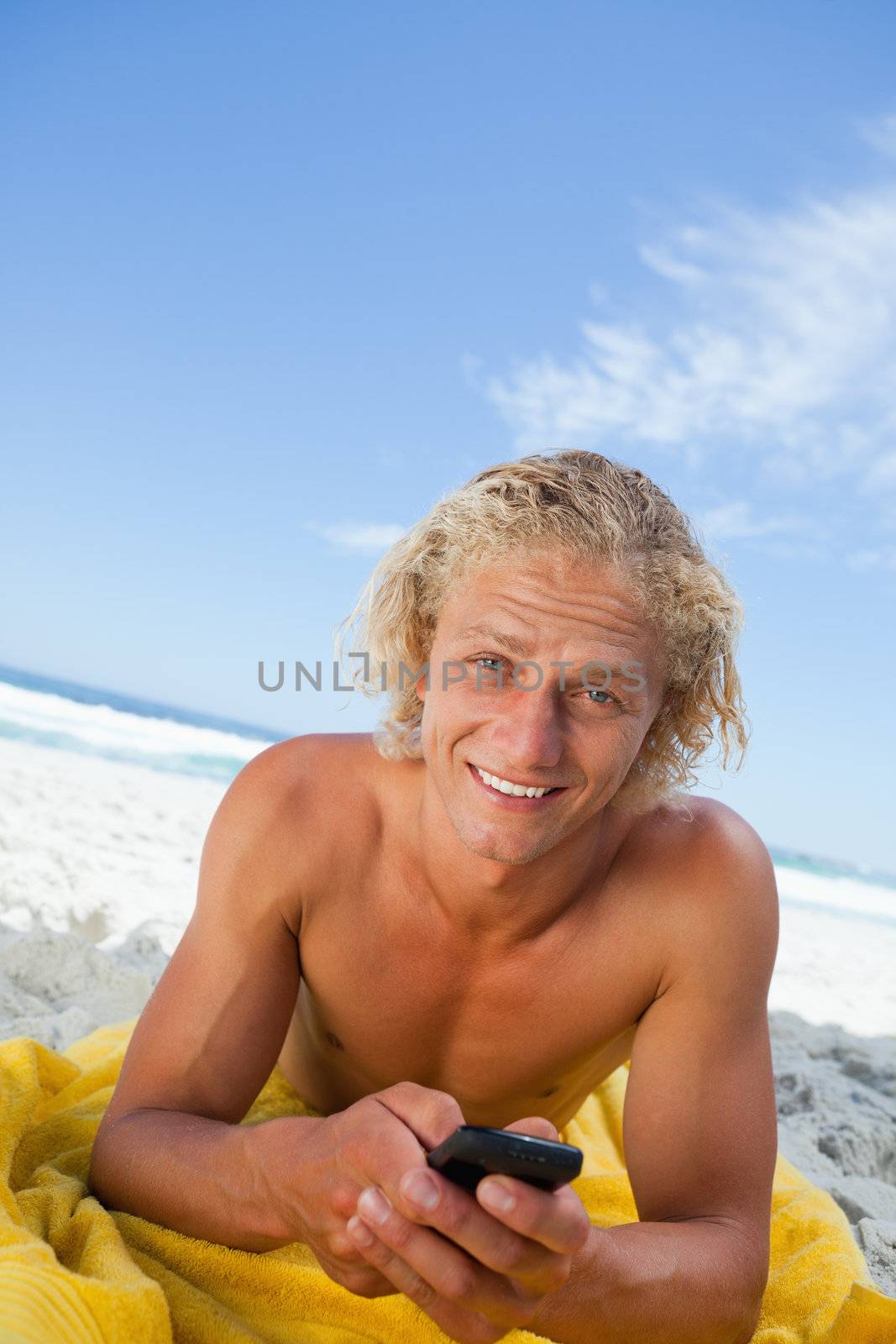Smiling blonde man sending a text while sunbathing and lying on his beach towel