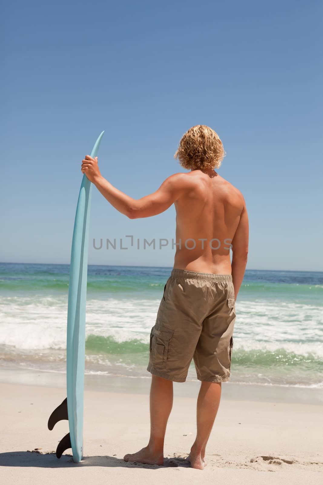 Young blonde man waiting for waves while holding his surfboard in front of the ocean