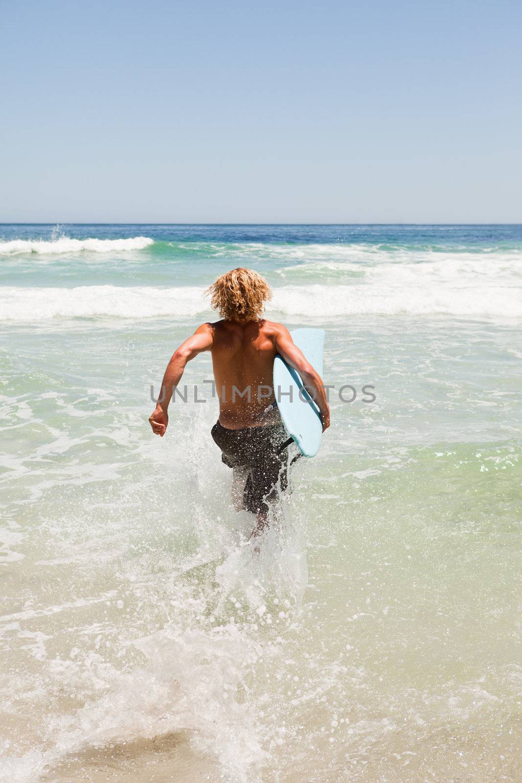 Young blonde man holding his blue surfboard while running in the water by Wavebreakmedia