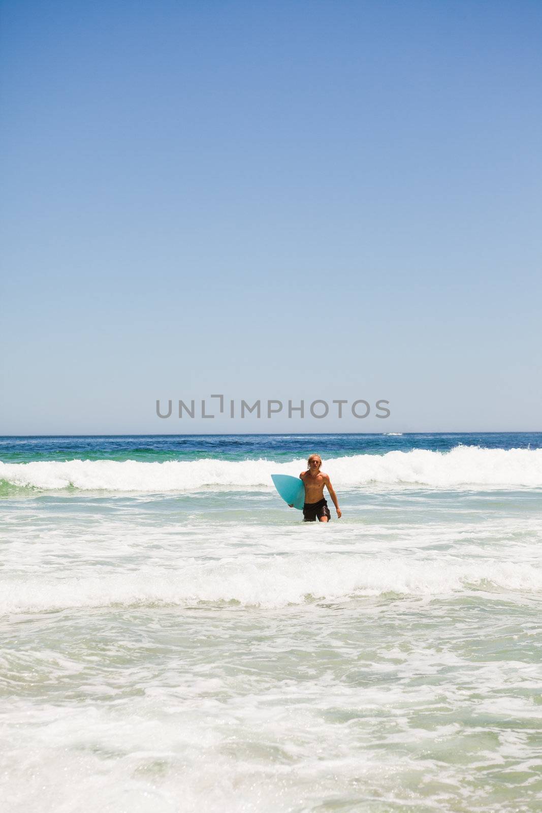 Young blonde man walking in the water while holding his blue surfboard