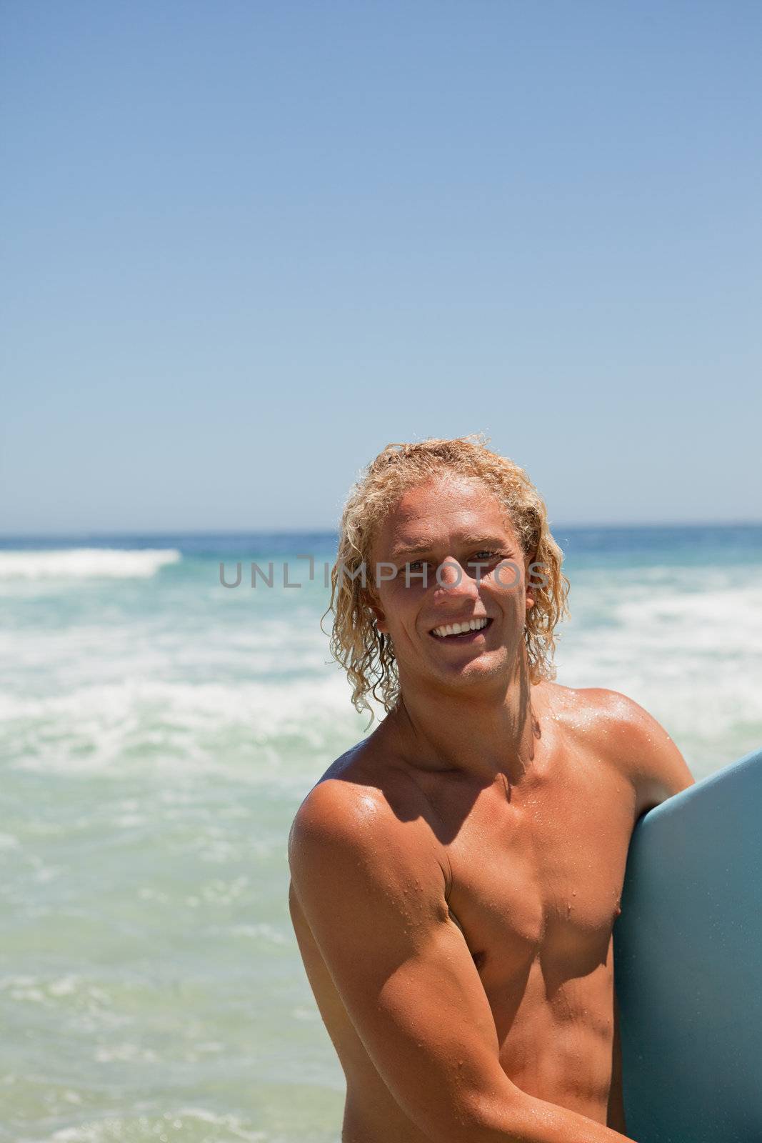 Young blonde man looking at the camera while smiling and holding his surfboard