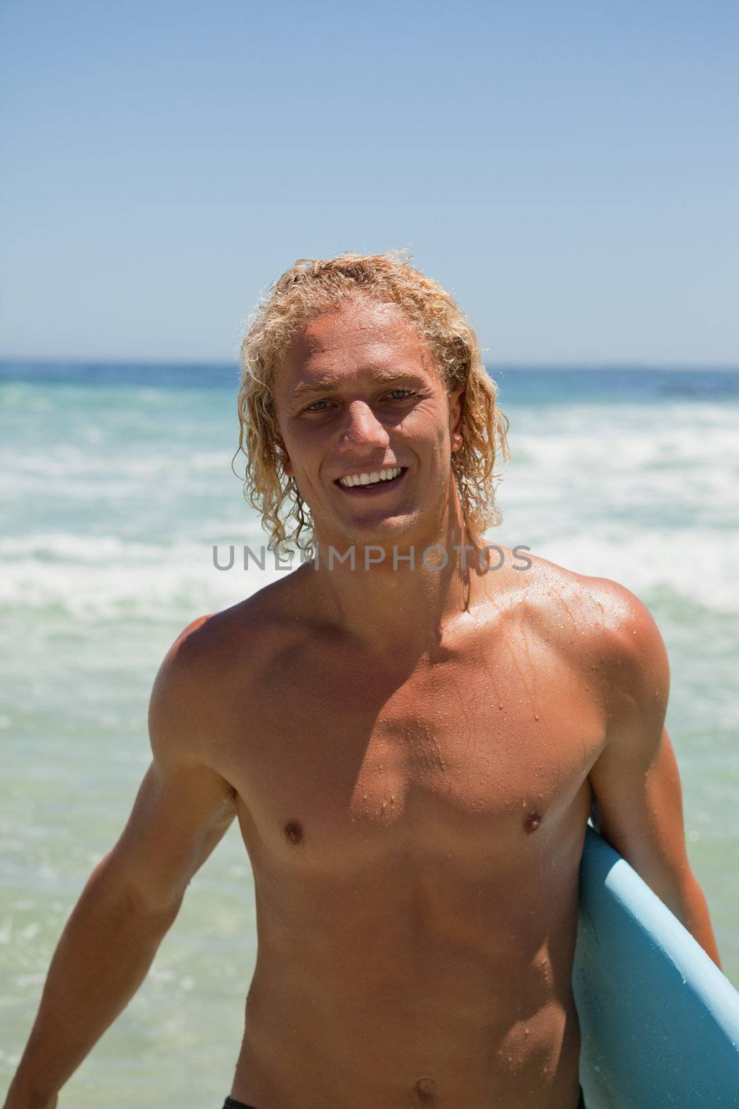 Smiling blonde man standing in the water with his blue surfboard