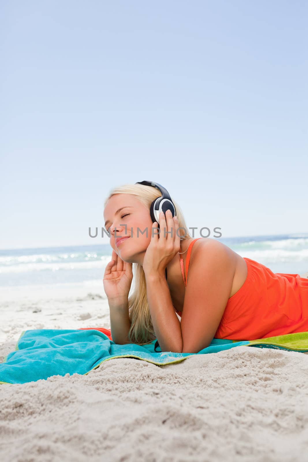 Thoughtful blonde woman lying on the beach while listening to music with her headset