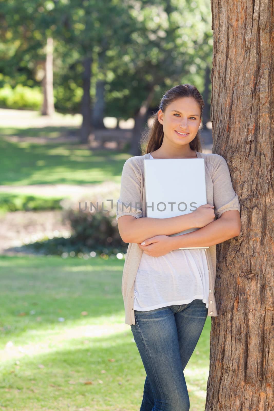 Smiling young woman holding a laptop while leaning against a tree