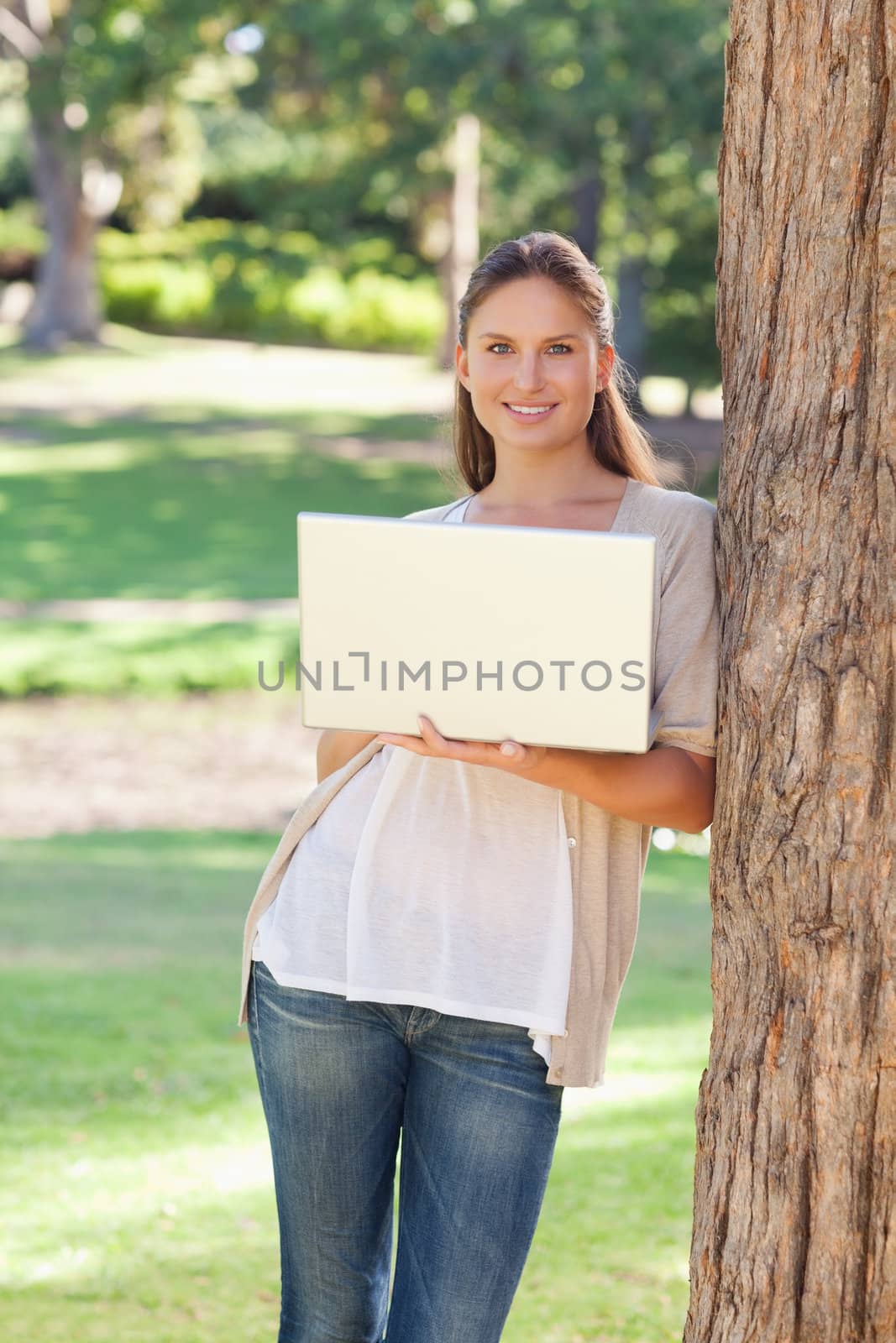 Smiling young woman using a laptop while leaning against a tree