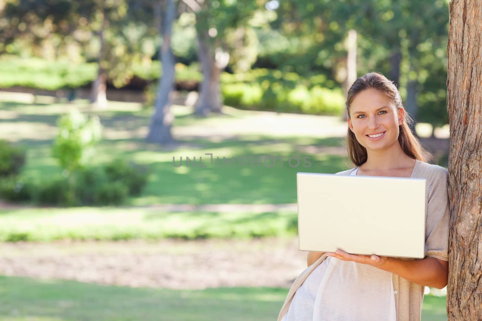 Smiling young woman using a notebook while leaning against a tree