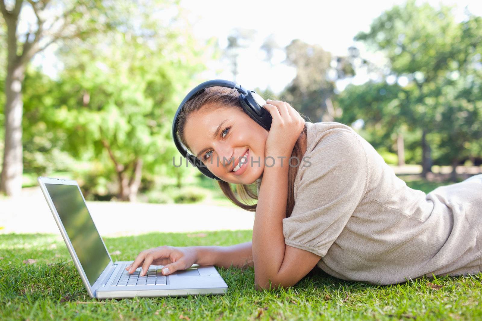 Side view of a smiling young woman with headphones and a laptop lying on the lawn