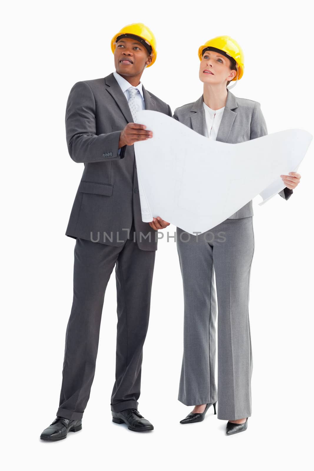 A businessman and woman wearing hard hats and holding a paper are looking up
