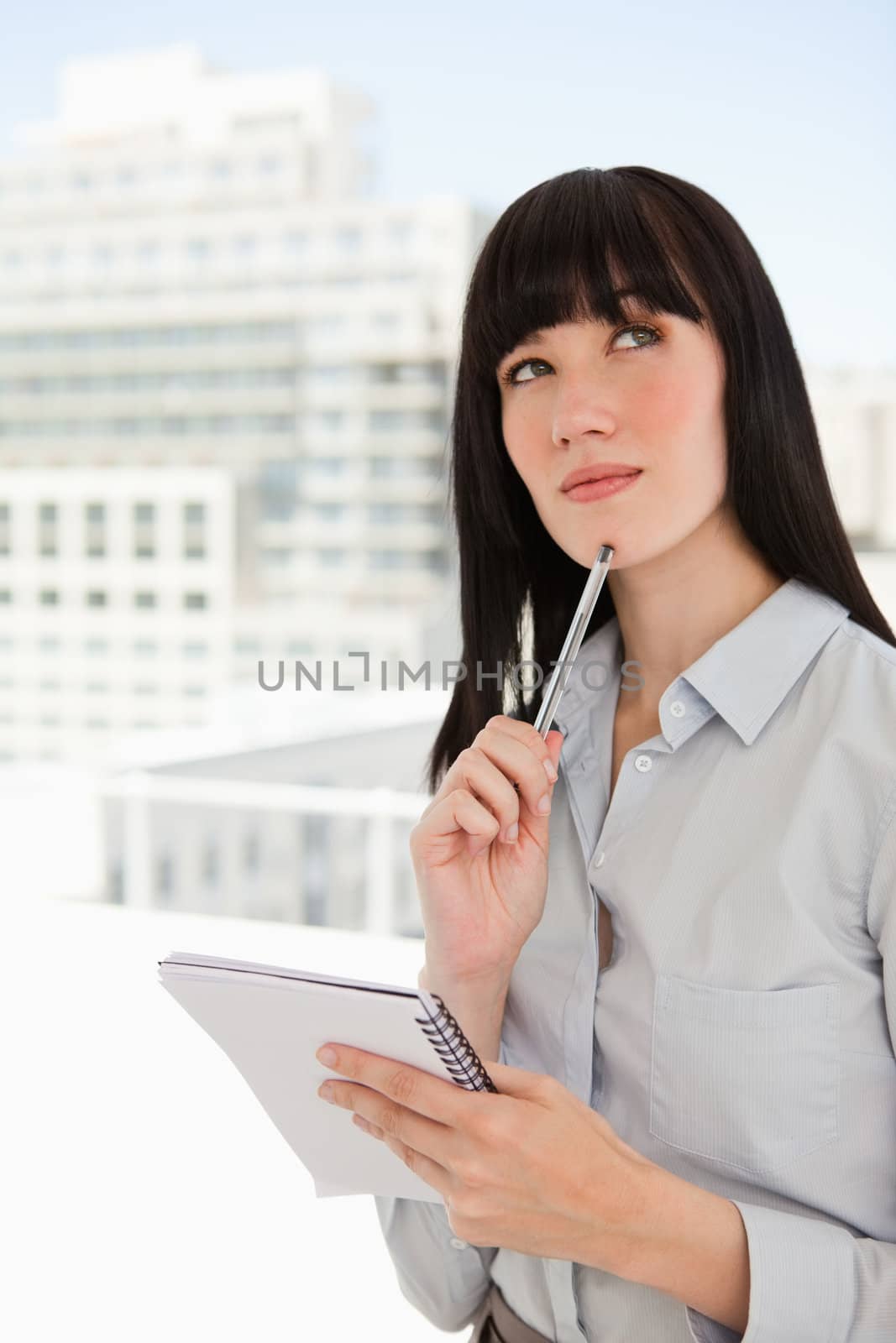 A woman in her office with a notepad and a pen against her chin looks upwards as she thinks