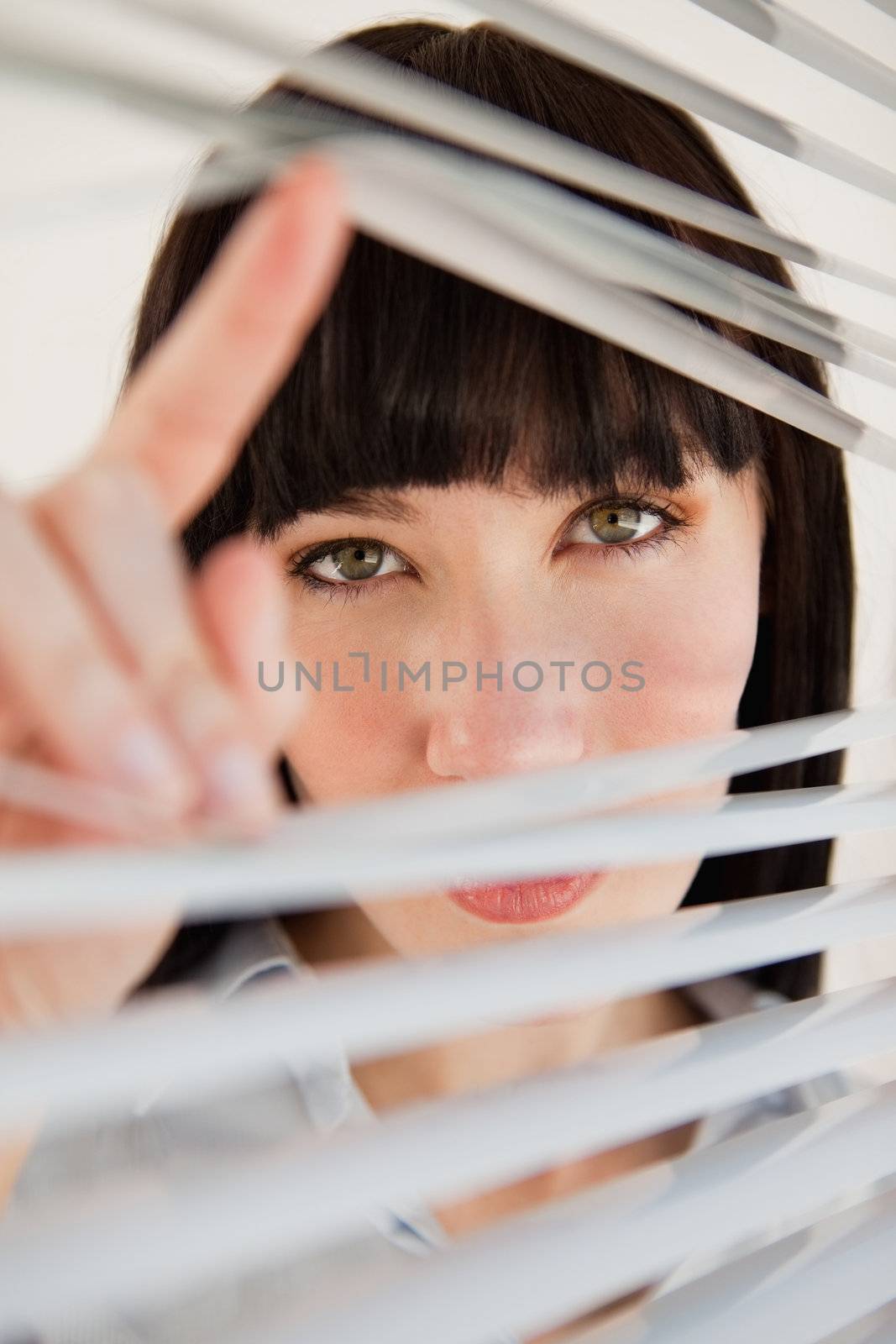A woman looking through some blinds into the camera by Wavebreakmedia