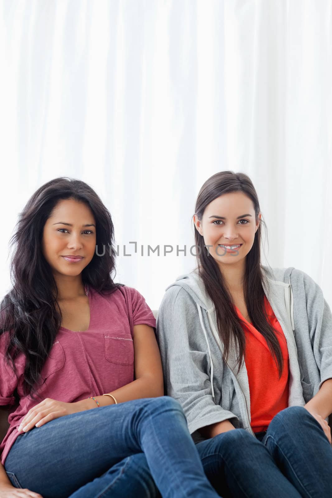 A half length-shot of two smiling women sitting on the couch while looking into the camera