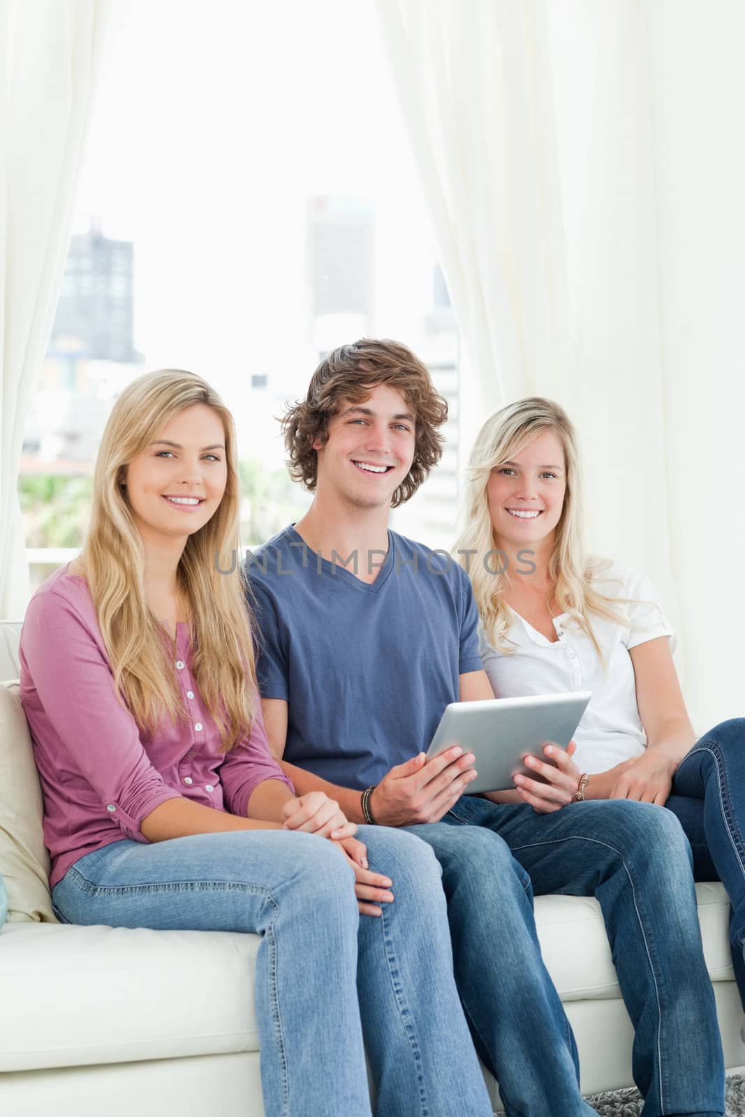 Two sisters and a brother sit together on the couch as the brother holds the tablet while they all look at the camera 