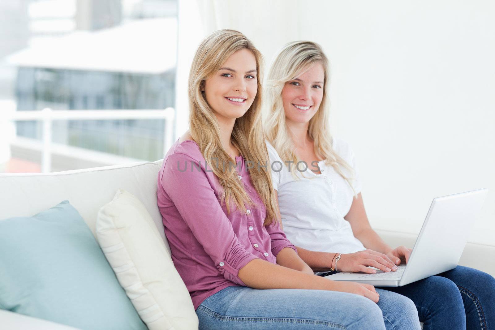 Smiling sisters sit on the couch holding a laptop while they look to the side into the camera 