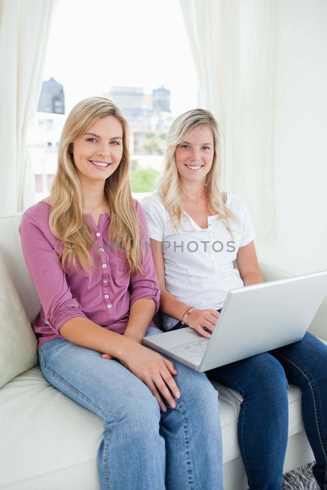 Sisters smiling as they sit on the couch together and hold a laptop while looking into the camera