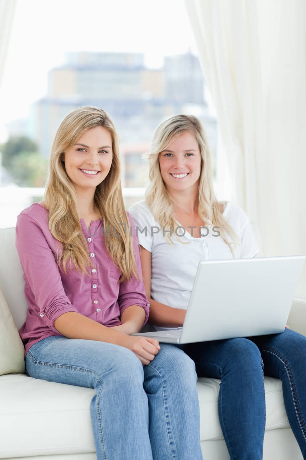 A pair of sisters sit together with a laptop on the couch as they look at the camera 