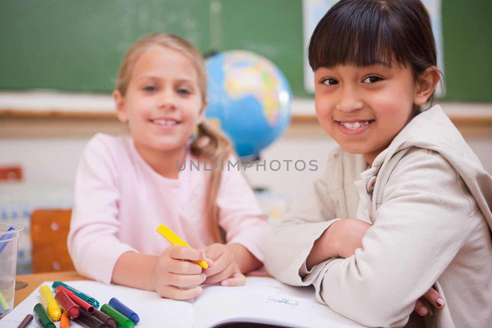 Smiling schoolgirls drawing while looking at the camera in a classroom