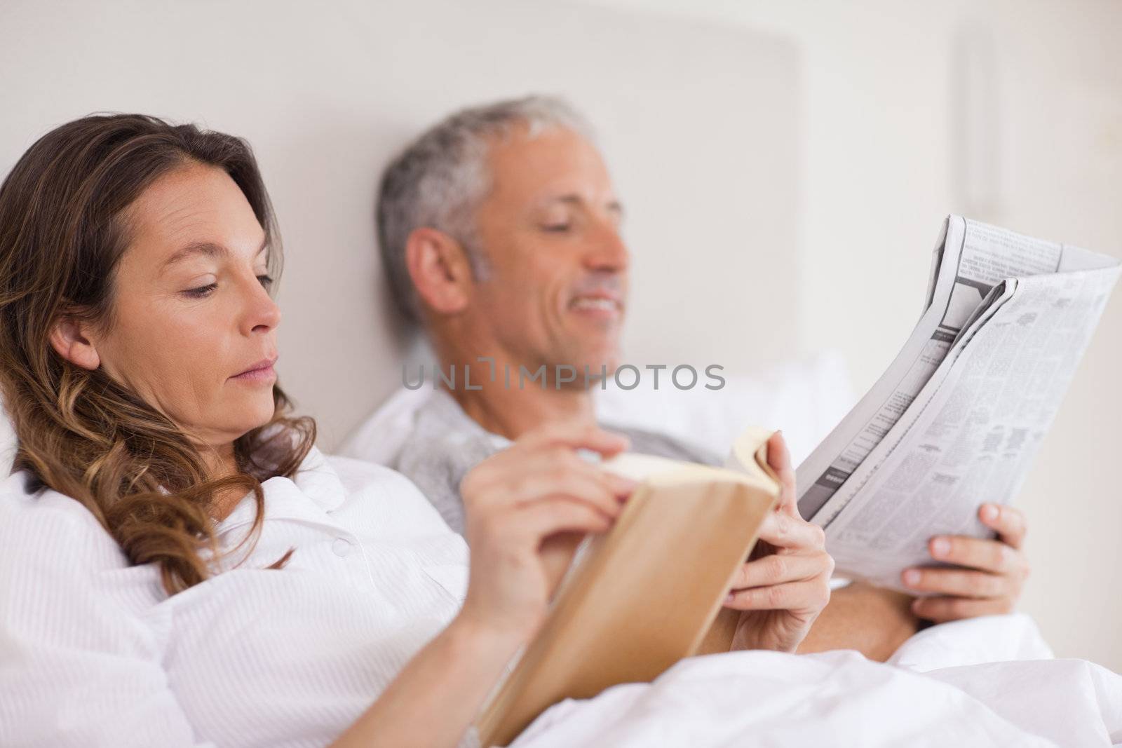 Woman reading a book while her husband is reading a newspaper in their bedroom