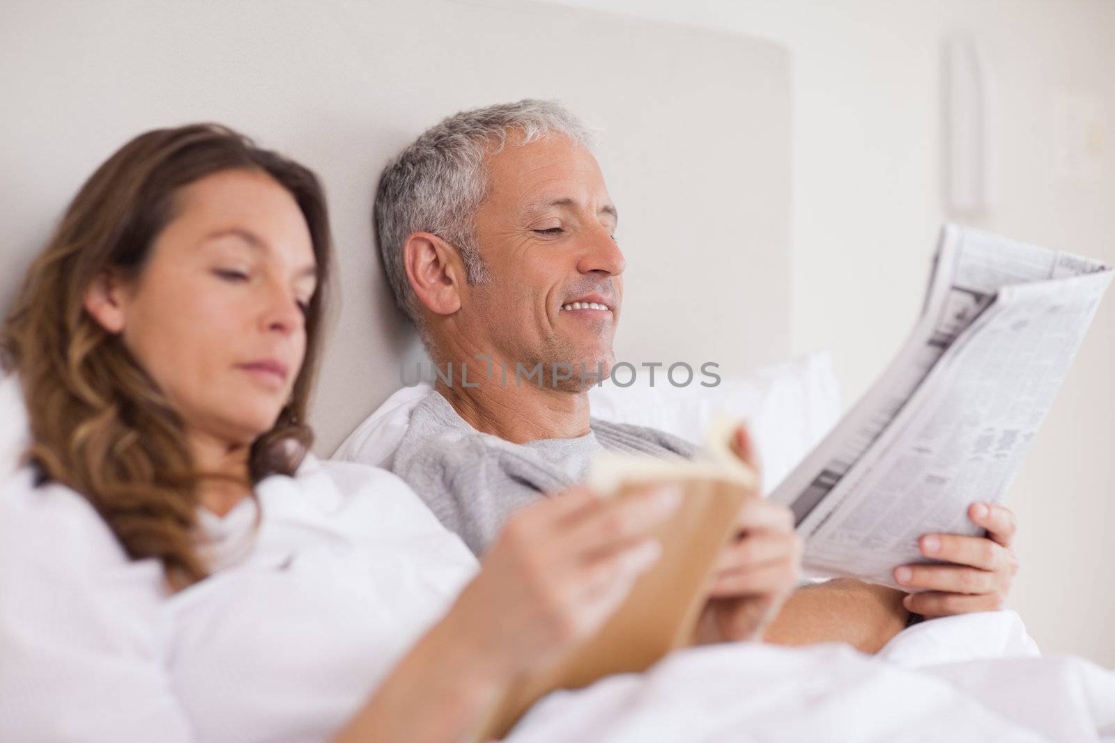 Happy woman reading a book while her husband is reading a newspaper in their bedroom