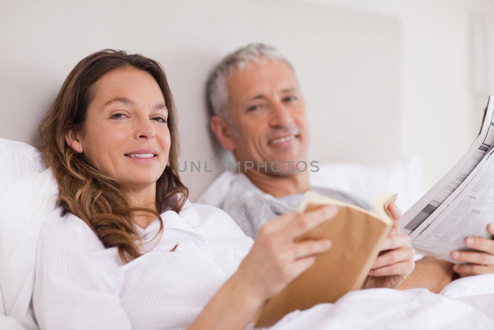 Smiling woman reading a book while her husband is reading the news in their bedroom