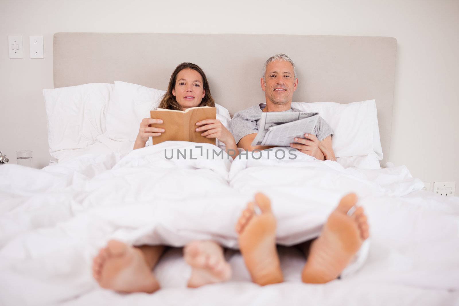 Man reading a newspaper while his wife is reading a book in their bedroom