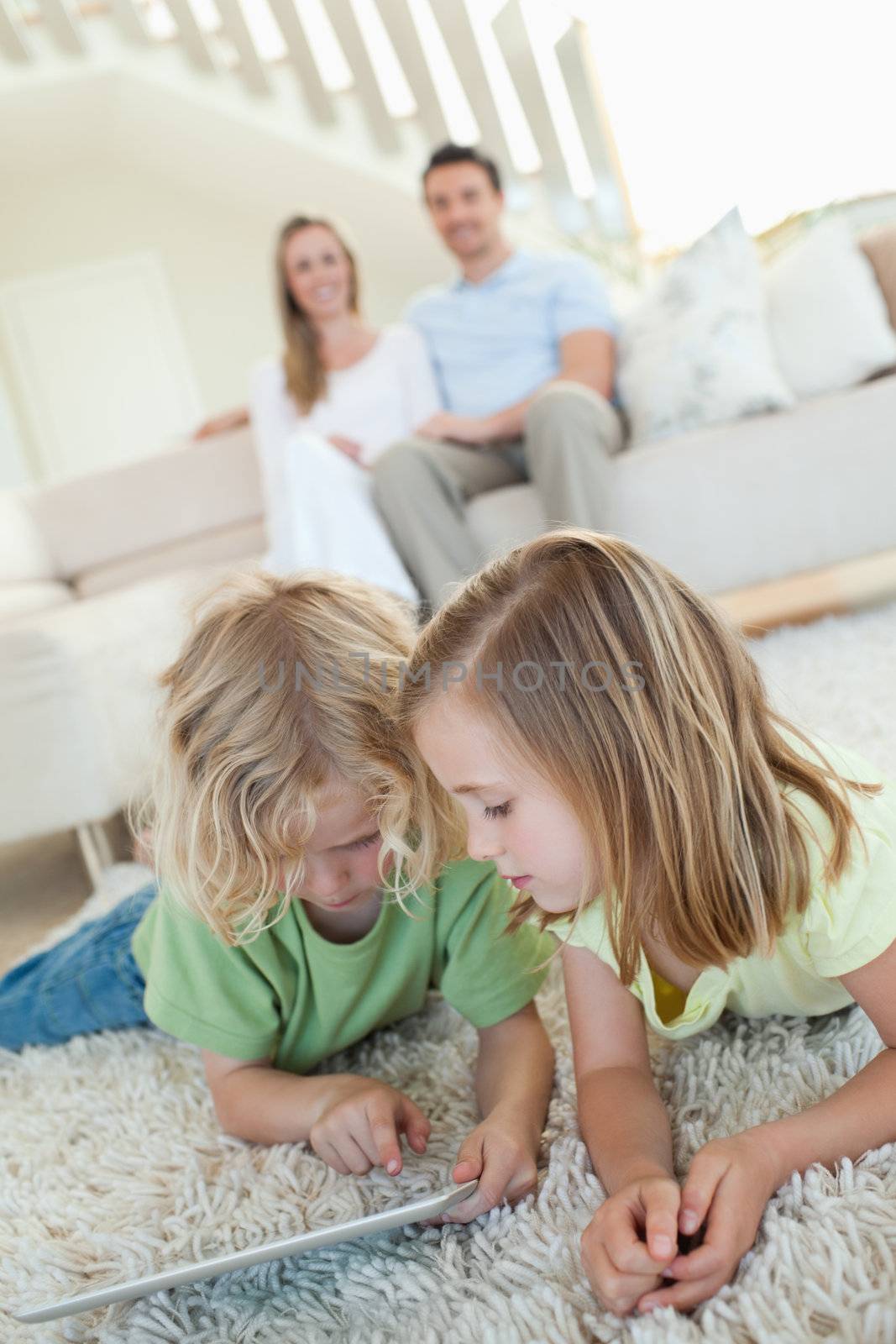 Siblings using tablet together on the carpet with parents behind them