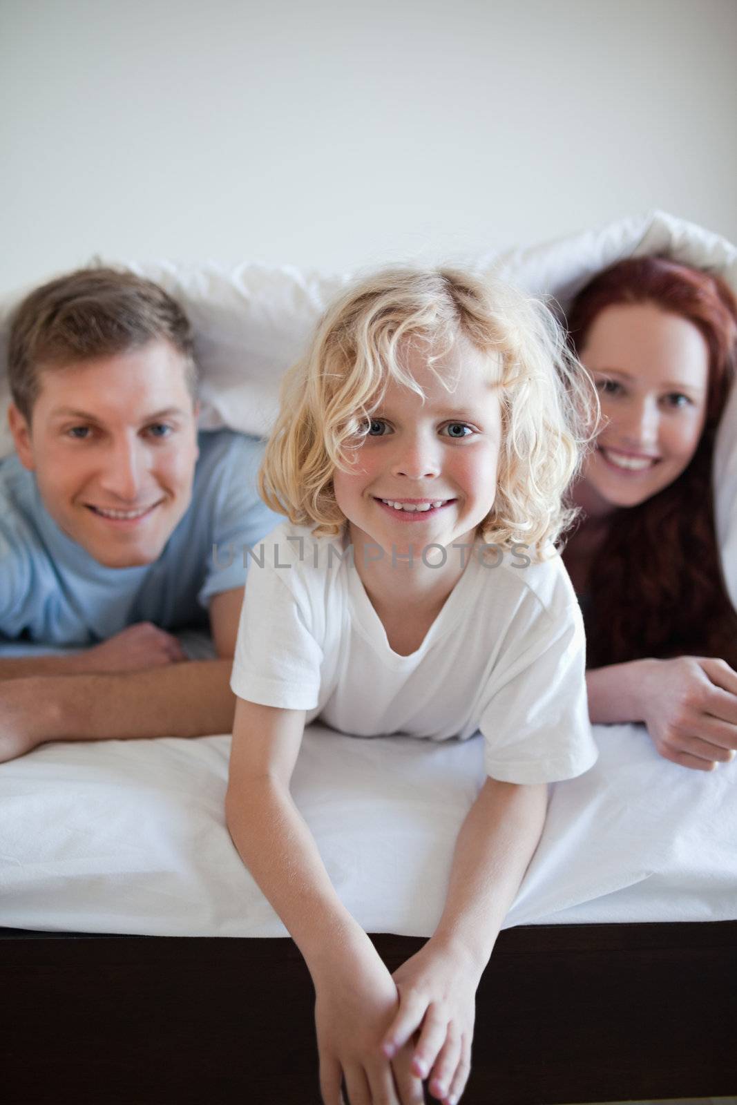 Cheerful boy under the bed cover together with his parents